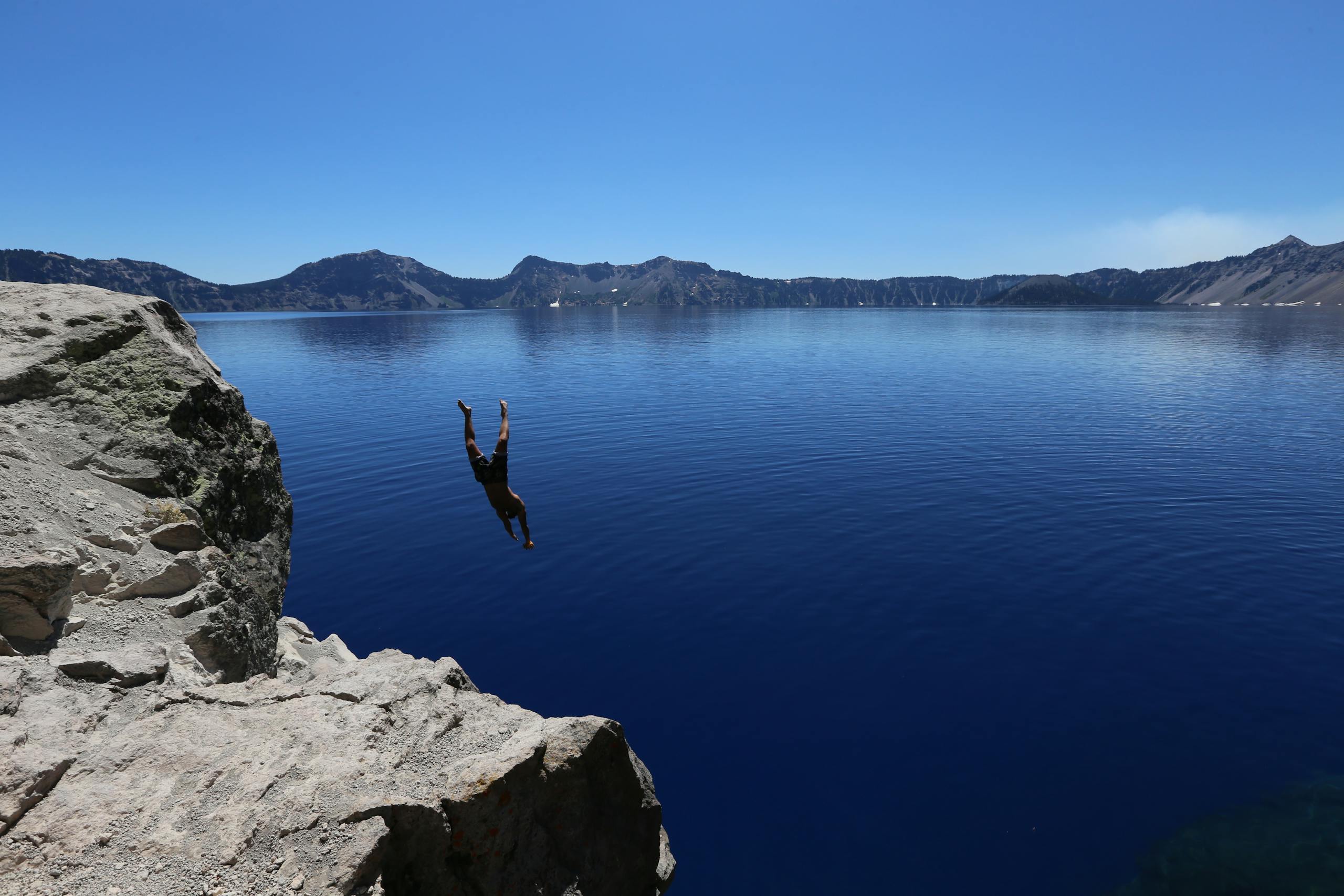 A person diving off a rocky cliff into Crater Lake's clear blue water under a bright sky.