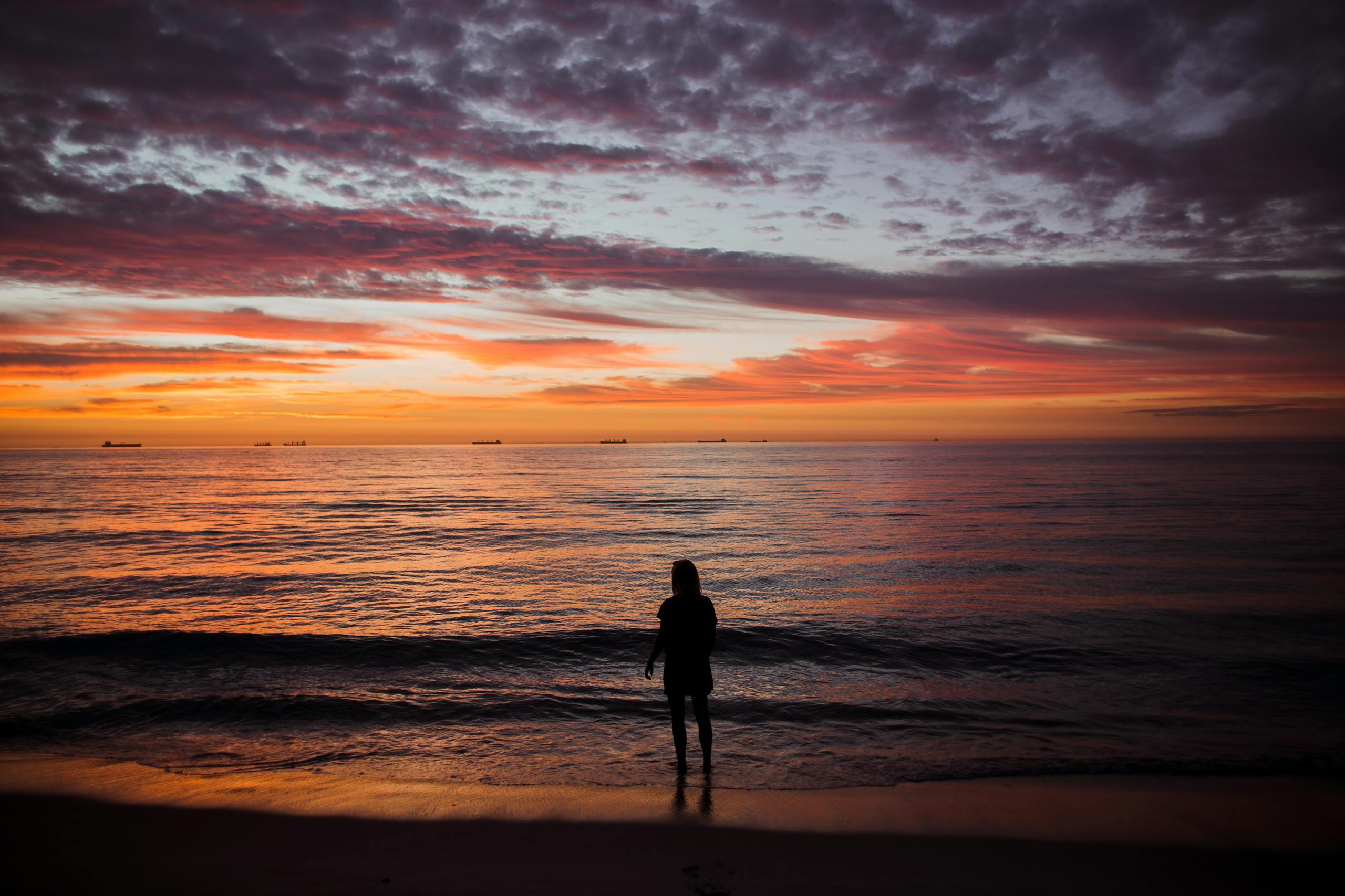 A woman stands silhouetted against a colorful sunrise over the tranquil beach and sea.
