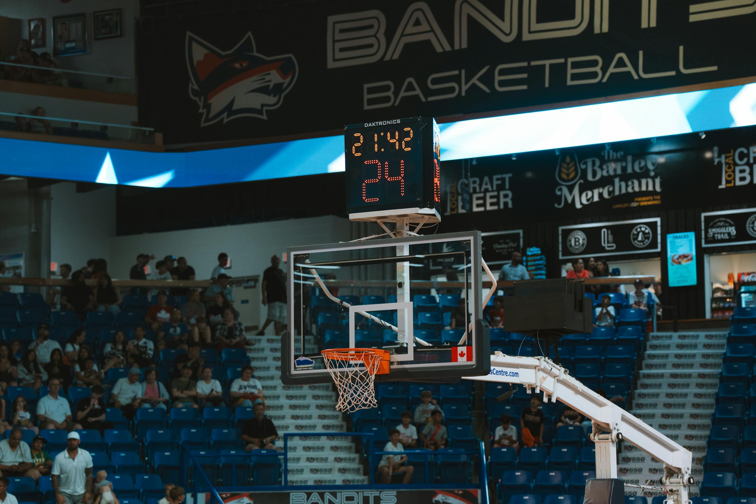 Close-up of a basketball hoop inside a stadium with a scoreboard and audience in the background.