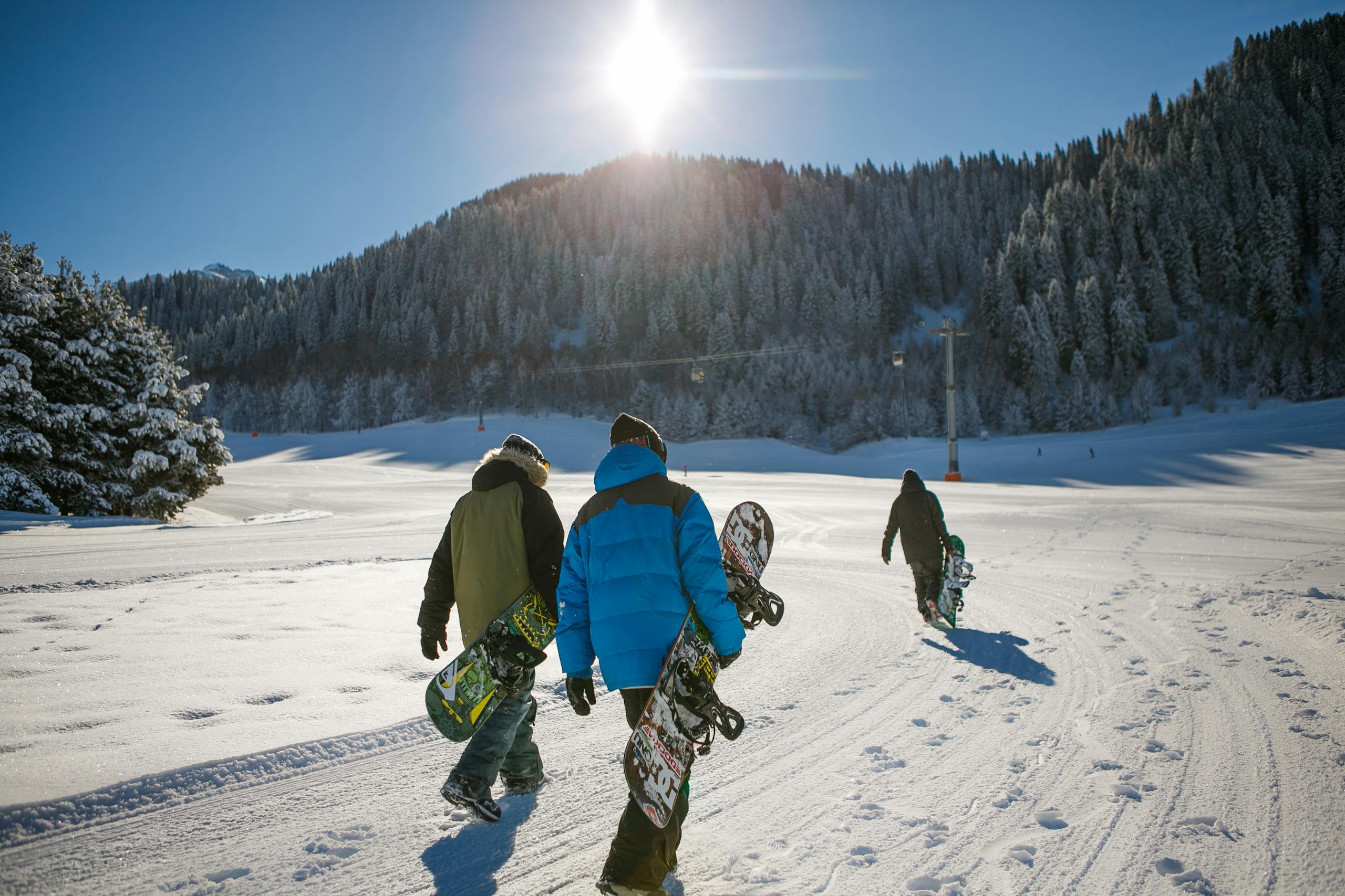 Three snowboarders walk on a snowy slope in bright winter sunlight, heading towards a scenic ski area.