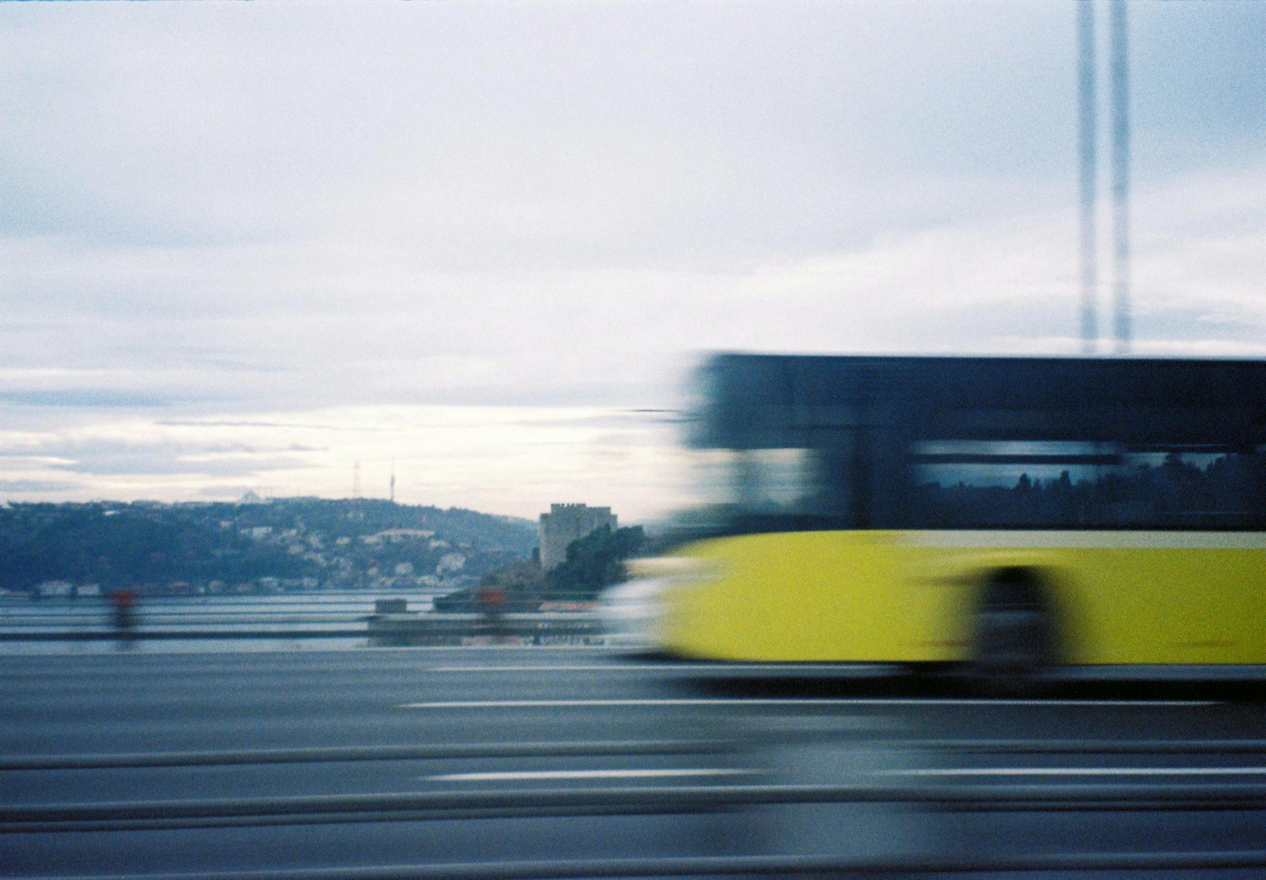 A blurred yellow bus speeds across a bridge with a scenic backdrop at dusk.