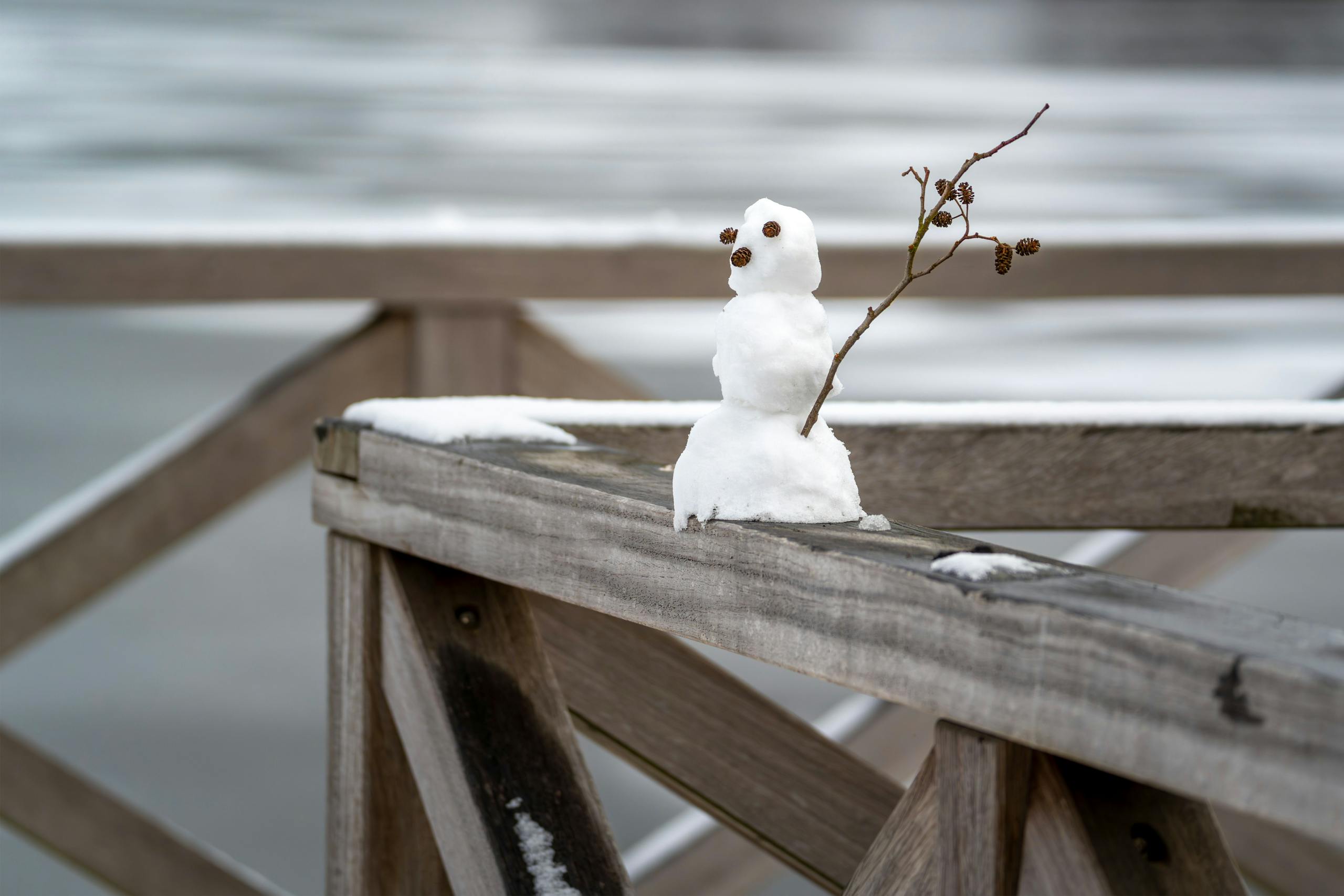 Miniature snowman on a snowy wooden fence by a frozen lake. Winter outdoor scene.