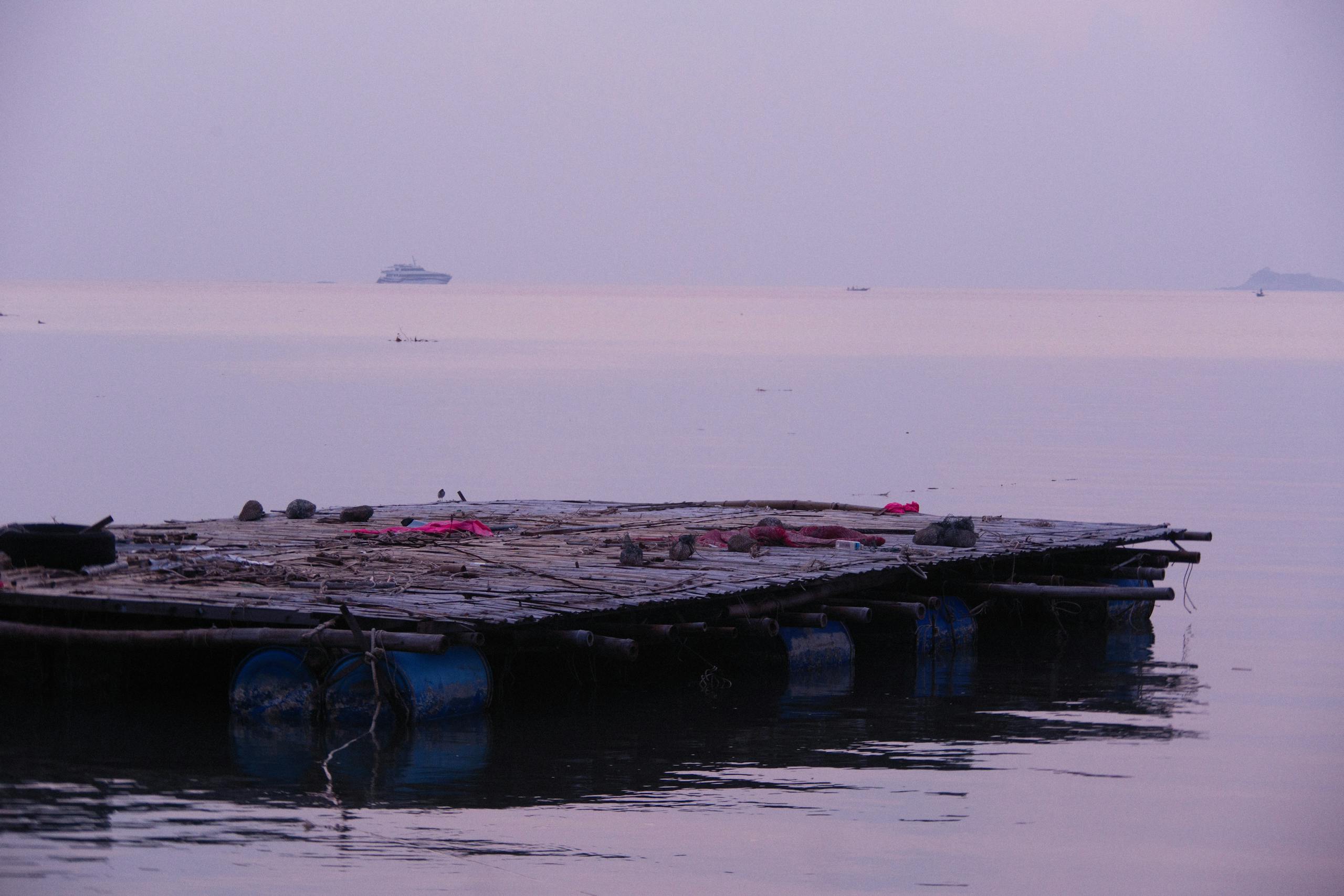 A handmade raft made from blue barrels floats on a calm sea with a boat in the distance under a soft, foggy sky.