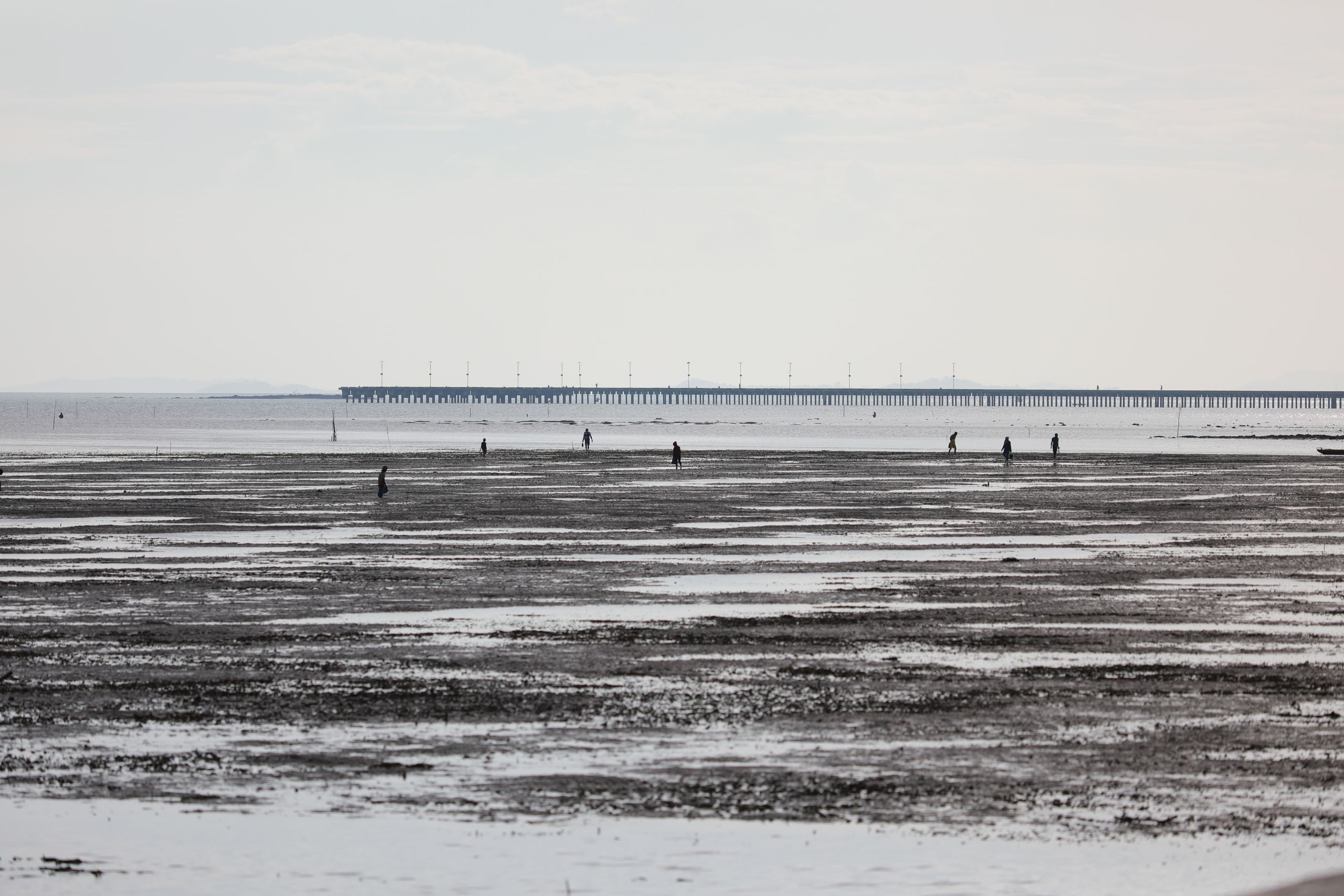 A tranquil beach at low tide with silhouettes of people and long pier in the distance under a soft sunset glow.