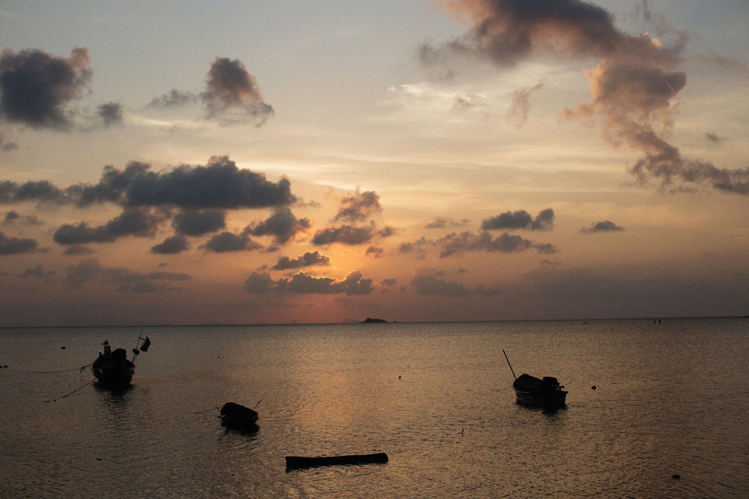 Peaceful sunset scene with silhouetted fishing boats and clouds reflecting on calm sea waters.