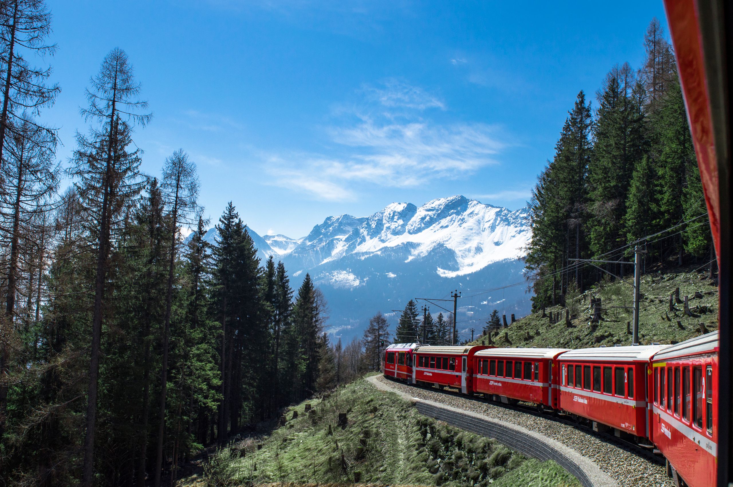A red train travels through lush forests and snowy mountains under a clear blue sky in Switzerland.